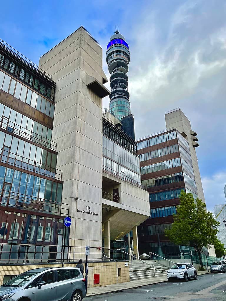 An urban college campus with multi-storey office buildings of brick, concrete, and glass. A tall blue tower rises behind the buildings.