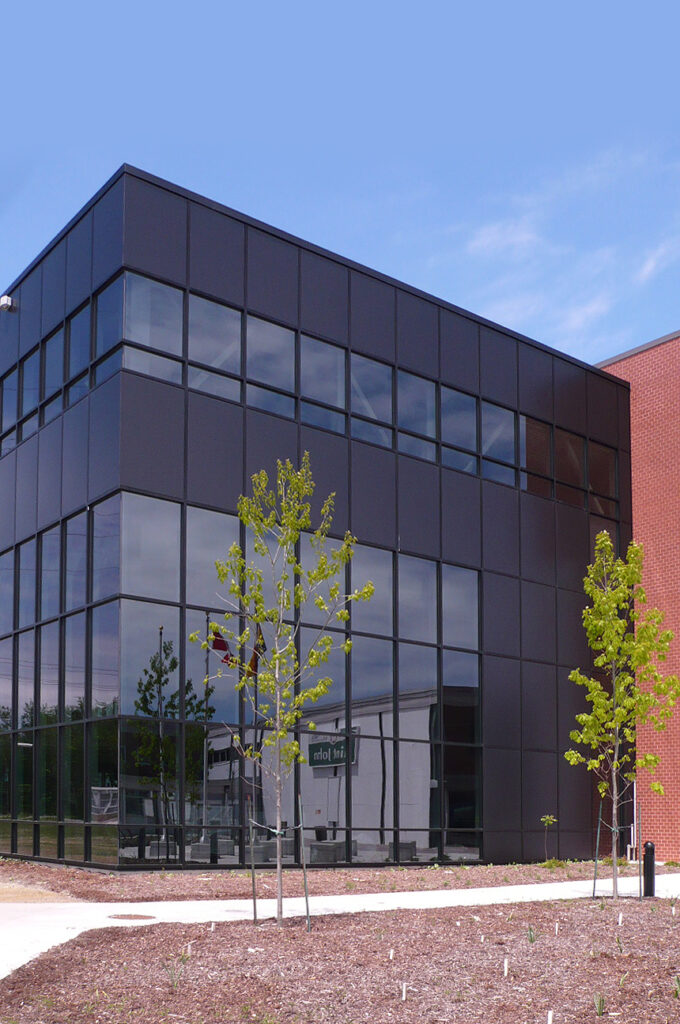 E-Building; a two-storey modern academic building, with young deciduous trees in a courtyard in front of it.