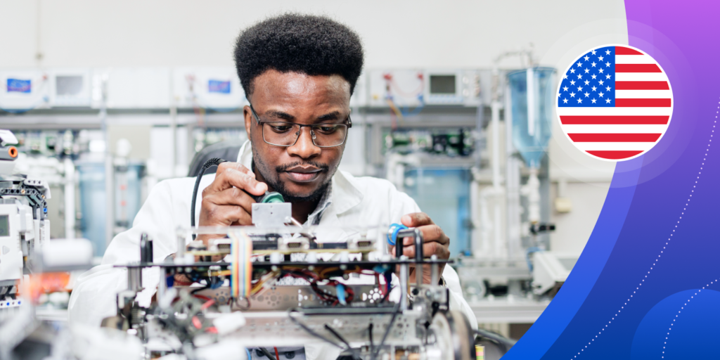 A young Black man works in an engineering lab environment, holding a soldering iron over a complex mechanical device.