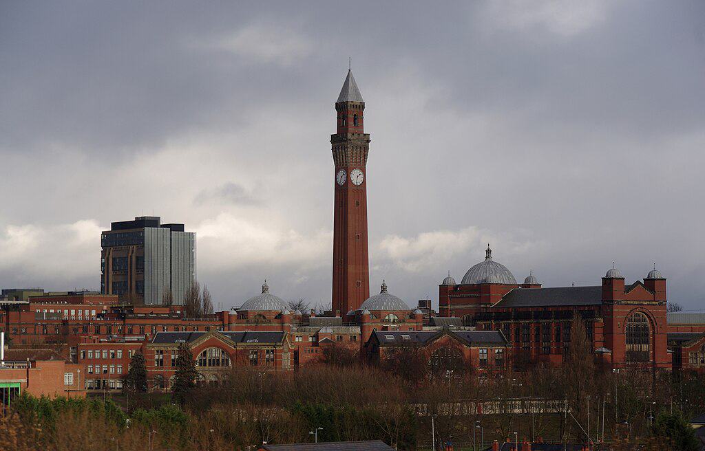 The University of Birmingham campus: multiple red-brick multistorey buildings arranged around a tall, red-brick clocktower. A more modern blue glass building is in the distance.