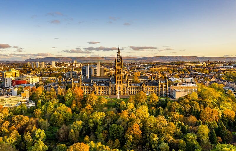 The University of Glasgow with Dumgoyne in the background (two-storey academic building with a tall central tower, fronted by deciduous trees in autumn foliage (greens, yellows, oranges) and a mid-sized city stretching out behind.