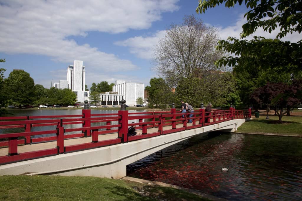 A red metal and beige concrete bridge spans a small creek in a city park in Huntsville, Alabama